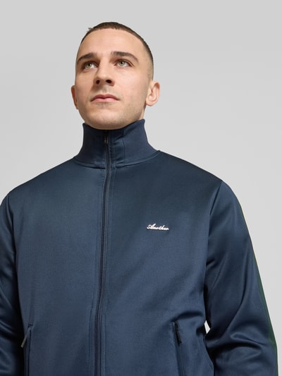 Männermodel trägt Track Jacket mit Stretch-Anteil Modell 'Everyday' von ANOTHER COTTON LAB, granatowy, elastyczna mieszanka materiałów, prosty krój, stójka, zamek błyskawiczny, kieszenie zapinane na zamek błyskawiczny, lampasy, aplikacja z metką, długość tyłu w rozmiarze M 63 cm, długość rękawów w rozmiarze M 68 cm, zbliżenie na górną część ciała