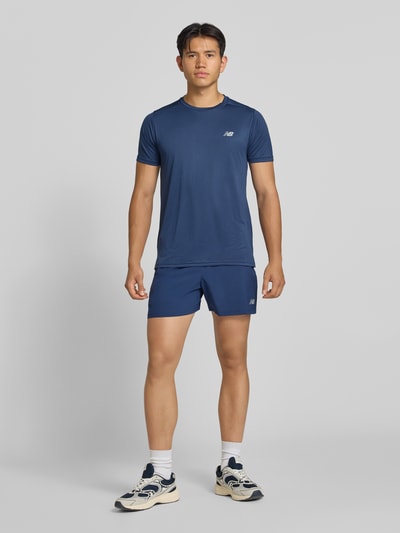 Männermodel trägt blaues Funktionsshirt mit Rundhalsausschnitt und Logo von New Balance, glatte Textur, Ganzkörperansicht von vorne, Regular Fit, Rückenlänge Größe M 69 cm.