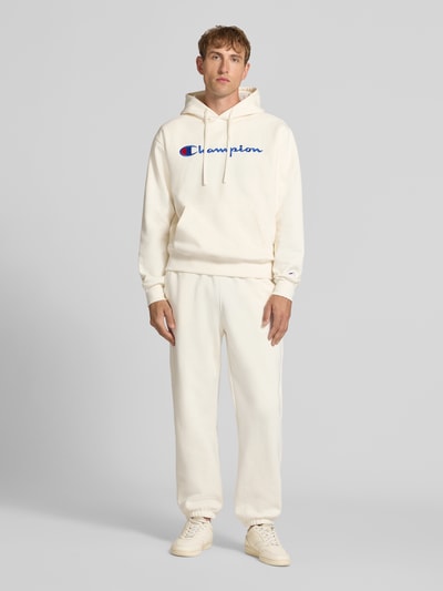 Männermodel trägt Hoodie mit Label-Stitching von Champion im Baumwoll-Mix, lockerer Schnitt, Kängurutasche, Ganzkörperansicht von vorne.