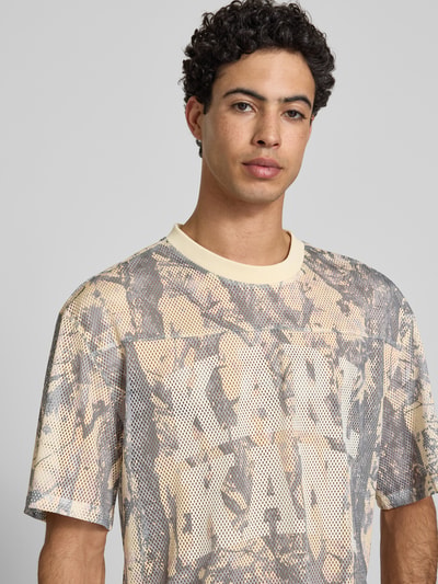 Männermodel trägt T-Shirt aus Mesh mit Logo von KARL KANI in Beige und Grau, Rundhalsausschnitt, Regular Fit, Nahaufnahme Oberkörper.