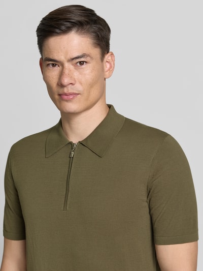 Męski model nosi Regular Fit koszulkę polo z krótkim zamkiem błyskawicznym model 'ORBIT' od Tiger Of Sweden z bawełny, krótki rękaw, kolor oliwkowy, długość tyłu w rozmiarze M: 68 cm, zbliżenie na górę torsu, widok z przodu