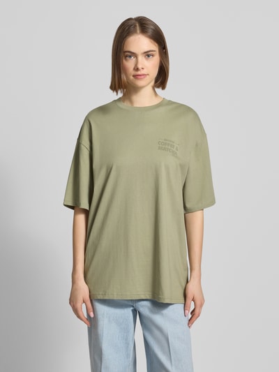 Modelka kobieca nosi Oversized T-Shirt mit Statement-Print od REVIEW, czysta bawełna, krój oversize, okrągły dekolt, jasna zieleń, nadruk z przodu, długość tyłu w rozmiarze S: 70 cm, widok całej sylwetki z przodu.