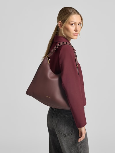 Kobiece model prezentuje torebkę z naturalnej skóry Handtasche aus echtem Leder Modell 'BOHEME' od Coccinelle w kolorze bordowym, ziarnista faktura, logo, pasek na ramię, Główna przegroda z zamkiem błyskawicznym, kieszeń wewnętrzna zapinana na zamek błyskawiczny, przegródki wewnętrzne, szerokość w rozmiarze 1: 30 cm, długość w rozmiarze 1: 32 cm, głębokość w rozmiarze 1: 12 cm, długość paska na ramię w rozmiarze 1: 44 cm, widok całej sylwetki z przodu.