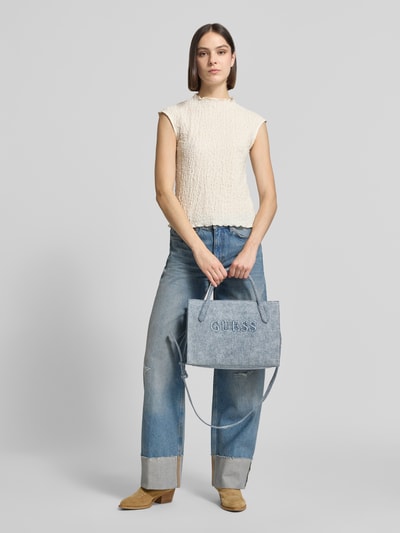 Frauenmodel trägt Tote Bag mit Viskose-Anteil Modell 'Jodi' von Guess in Denim-Optik mit Logo-Detail, Hauptfach mit Reißverschluss, Baumwoll-Viskose-Mix, Schulterriemen, abnehmbare Trageriemen, rechteckige Form und strukturierte Oberfläche, Breite 34 cm, Länge 24 cm, Tiefe 12 cm, Länge Schulterriemen 112 cm, Ganzkörperansicht von vorne.