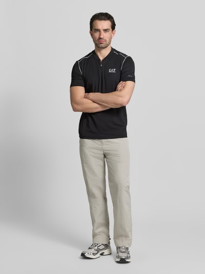 Männermodel trägt Regular Fit T-Shirt mit Stehkragen von EA7 Emporio Armani, Schwarz mit Label-Print, Ganzkörperansicht von vorne.