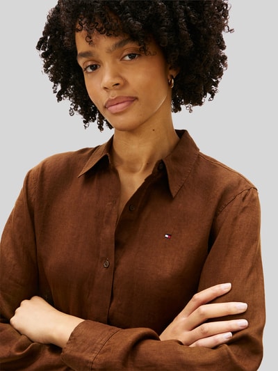 Frauenmodel trägt Relaxed Fit Langarmbluse aus reinem Leinen von Tommy Hilfiger, Optik: Unifarben, Passform: Relaxed-Fit, Hemdkragen, Logo-Stitching, Verschluss: Knopfleiste, Ärmelform: Langarm, Ärmelabschluss: Manschette, Rumpfabschluss: abgerundet, Materialverarbeitung: Flammgarn, Material: Leinen, Textur wirkt matt, warme Braunfarbe, Nahaufnahme Oberkörper.