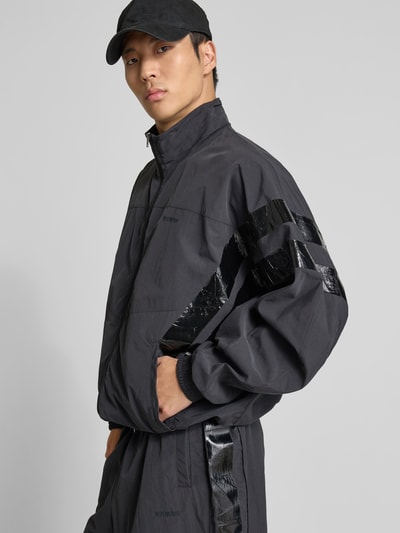 Männermodel trägt schwarze Trainingsjacke mit Stehkragen von VETEMENTS, Relaxed-Fit, Zweiwege-Reißverschluss, elastische Abschlüsse, Eingrifftaschen, glatte Textur, Rückenlänge 74 cm, Schulterbreite 50 cm, Ärmellänge 73 cm, Ganzkörperansicht von vorne.
