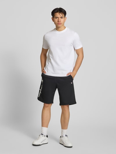 Männermodel trägt schwarze Shorts mit Logo-Print und -Stitching von CHAMPION, lockerer Schnitt, Baumwoll-Mix, Ganzkörperansicht von vorne.