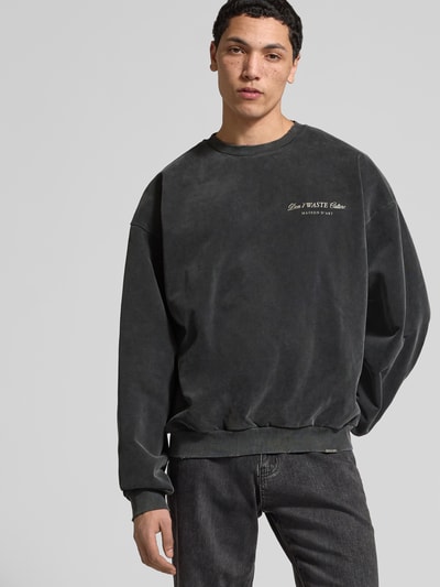 Männermodel trägt Oversized Sweatshirt aus Baumwoll-Mix von Don't Waste Culture in Dunkelgrau mit Statement-Print, elastische Abschlüsse, Nahaufnahme Oberkörper. Rückenlänge bei Größe M: 67 cm.