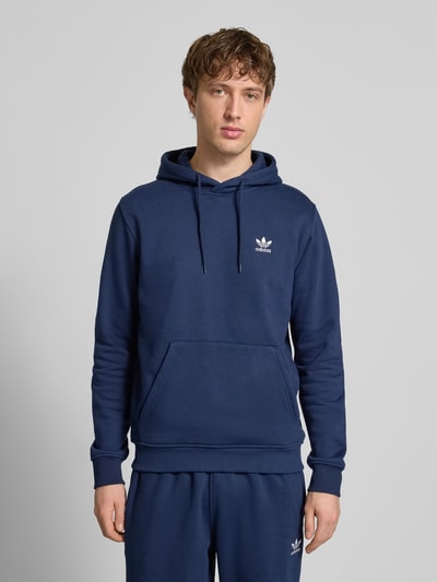 Męski model nosi Hoodie mit Kapuze od adidas Originals w kolorze granatowym, bawełniany miks, luźny krój, z kapturem, tunel, label-stitching, kieszeń kangurka, ściągacze, długość tyłu w rozmiarze M: 68 cm, długość rękawa: 69 cm, widok całej sylwetki z przodu.