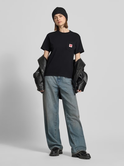 Kobiece model prezentuje damski t-shirt z bawełny o prostym kroju z okrągłym dekoltem i brustaszą z naszywką z logo Carhartt Work In Progress, długość tyłu 58 cm w rozmiarze S, kolor czarny, gładka tekstura, całkowity widok z przodu.