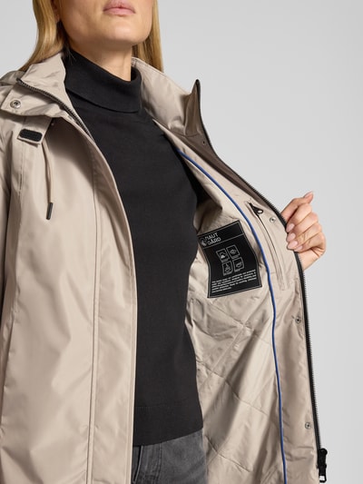 Frauenmodel trägt Regular Fit Mantel mit Logo-Applikation von NAUTGÅRD in Beige mit Kapuze, Stehkragen und Eingrifftaschen, Nahaufnahme Oberkörper. Rückenlänge 88 cm, Schulterbreite 37 cm, Brustweite 48 cm, Ärmellänge 63 cm, kontrastierendes Futter.