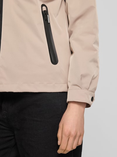 Kobiece modelka nosi kurtka z stójką Jacke mit Stehkragen od khujo, krój regular fit, zamek błyskawiczny, kieszenie zapinane na zamek błyskawiczny, długość tyłu w rozmiarze S: 60 cm, szerokość ramion w rozmiarze S: 51 cm, szerokość klatki piersiowej w rozmiarze S: 50 cm, długość rękawów w rozmiarze S: 54 cm, jasny beż, czarna kieszeń, zbliżenie na górę ciała