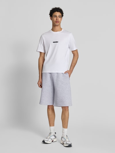 Mężczyzna nosi Sweatshorts mit Logo-Print od REVIEW, mieszanka bawełny, krój loose fit, elastyczny pas, nadruk z logo, jasnoszary kolor, szerokość w rozmiarze M 45 cm, długość wewnętrzna nogawki w rozmiarze M 28 cm, zdjęcie całej sylwetki z przodu.
