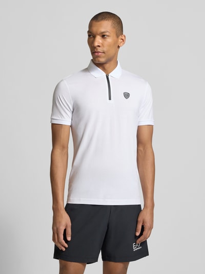 Męski model nosi Slim Fit Poloshirt aus Viskose-Mix von EA7 Emporio Armani, biały, detal z logo, kołnierzyk polo, elastyczne zakończenia rękawów, długość tyłu w rozmiarze M: 60 cm, mieszanka wiskozy, krój slim fit, Ganzkörperansicht z przodu.