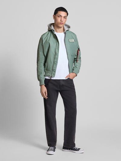 Männermodel trägt blouson mit logohanger Modell 'MA-1 TT' von Alpha Industries in groen met grijze capuchon, los model, labelpatch, mouwzak met logohanger, ritssluiting, elastische boorden; ruglengte maat M: 69 cm, schouderbreedte: 46 cm, borstwijdte: 54 cm, mouwlengte: 67 cm, ganzkörperansicht von vorne