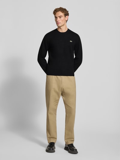 Männermodel trägt Regular Fit Pullover aus reiner Wolle von Lacoste in Schwarz, kombiniert mit beiger Hose, Ganzkörperansicht von vorne. Herren Pullover von Lacoste