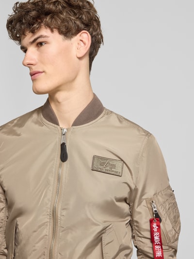 Männermodel trägt Regular Fit Bomberjacke mit Logo-Patch von Alpha Industries in Beige, glänzende Textur, Nahaufnahme Oberkörper.