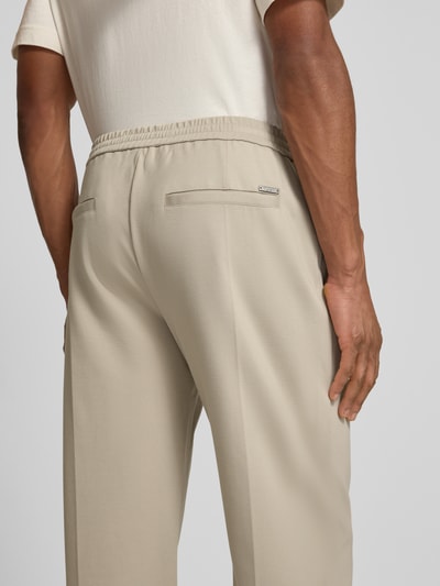 Männermodel trägt Slim Fit Hose aus Viskose-Mix Modell 'ALTON' von Guess in Beige mit Stretch-Anteil, Bundfalten, Eingrifftaschen, Ganzkörperansicht Rückansicht.