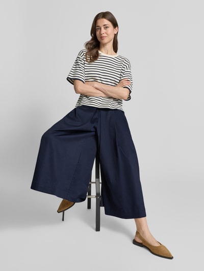 Vrouwenmodel draagt Wide Fit stoffen broek met bandplooien van Christian Berg Woman, puur katoen, verkorte pasvorm, elastische band, donkere blauwe kleur, binnenbeenlengte maat 36: 61 cm, bandwijdte maat 36: 36 cm. Volledige lichaamsweergave vooraan.