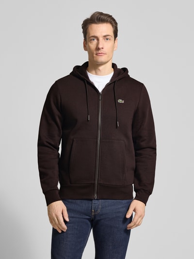 Męski model nosi Regular Fit Sweatjacke aus Baumwoll-Mix von Lacoste, jednokolorowy, mieszanka bawełny, krój regular fit, zamek błyskawiczny, naszywka z logo, kaptur z tunelem, długi rękaw, prążkowane zakończenia, przednie kieszenie, dzianina dresowa. Ganzkörperansicht von vorne.