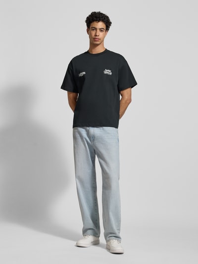 Männermodel trägt T-Shirt mit Label-Print von Another Cotton Lab, Reine Baumwolle, Lockerer-Schnitt, schwarze Farbe, Ganzkörperansicht von vorne.