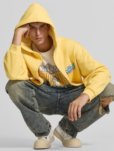 Męski model nosi bluzę rozpinaną 'Sweatjacke aus reiner Baumwolle' od RHUDE, bawełna, krój relaxed fit, kaptur, kieszeń kangurka, zamek błyskawiczny dwustronny, długość tyłu w rozmiarze M: 60 cm, szerokość ramion w rozmiarze M: 64 cm, długość rękawów w rozmiarze M: 54 cm, jasny żółty, tekstura gładka, widok z przodu, ujęcie w pełnej długości.