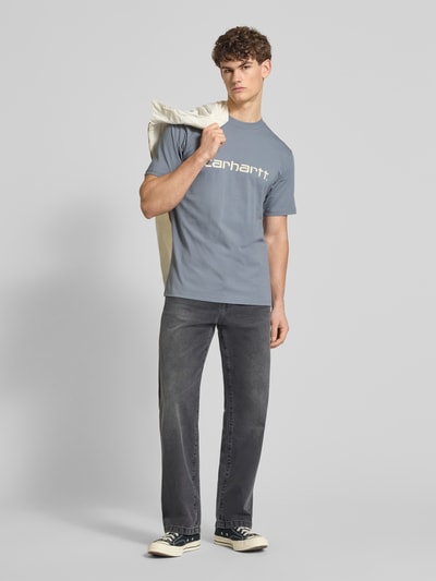 Männermodel trägt T-Shirt aus Baumwolle Modell 'Script' von Carhartt Work In Progress, gerader Schnitt, Rundhalsausschnitt, Label-Print, Ganzkörperansicht von vorne.