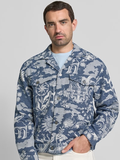 Męski model trägt kurtkę 'Jacke im Allover-Look mit Reverskragen' von Scotch & Soda, krój regular fit, wzór na całej powierzchni, kołnierz z połami, listwa guzikowa, kieszenie na piersi, długość tyłu w rozmiarze M: 61 cm, szerokość ramion w rozmiarze M: 43 cm, szerokość klatki piersiowej w rozmiarze M: 51 cm, długość rękawów w rozmiarze M: 62 cm, niebieskie i białe odcienie, tekstura tkanej tkaniny, całkowity widok z przodu.