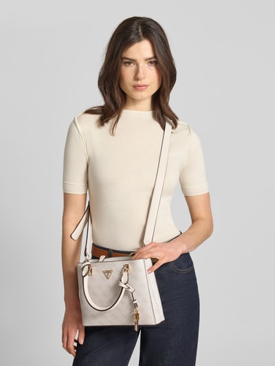 Frauenmodel trägt Handtasche mit Logo-Muster Modell 'NOELLE II' von Guess in Beige mit Applikation, Hauptfach mit Reißverschluss, Nahaufnahme Oberkörper.