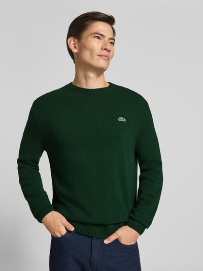Männermodel trägt Regular Fit Pullover aus reiner Wolle von Lacoste in Dunkelgrün, feine Textur, Ganzkörperansicht von vorne. Herren Pullover von Lacoste