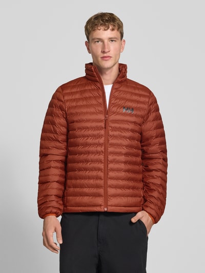 Männermodel trägt Steppjacke mit Label-Print von Helly Hansen in Rostrot, Stehkragen, durchgehender Reißverschluss, Ganzkörperansicht von vorne, gerade Passform, elastische Ärmelabschlüsse, sichtbarer Label-Print.