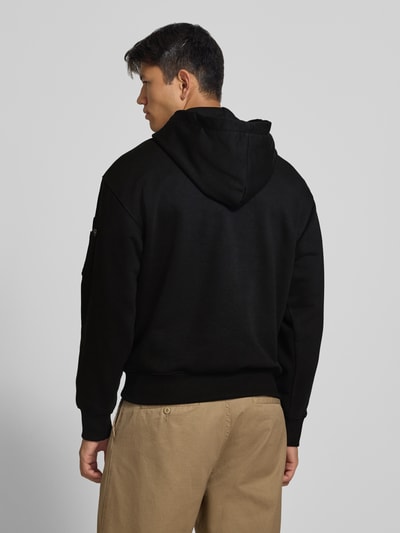 Männermodel trägt Hoodie mit Label-Print von Alpha Industries in Schwarz, Baumwoll-Mix, lockerer Schnitt, Rückansicht, Ganzkörperansicht.