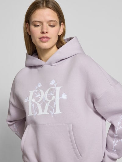 Frauenmodel trägt Relaxed Fit Hoodie mit Logo-Print von Review in Flieder, florales Stitching, Känguru-Tasche, Nahaufnahme Oberkörper.