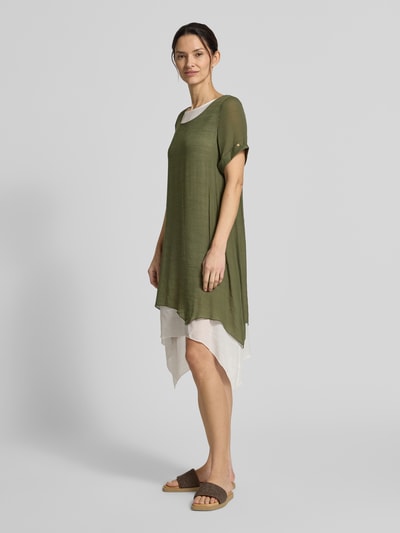 Frauenmodel trägt Knielanges Kleid aus Viskose-Mix von Apricot in Olivgrün mit weißem Unterkleid, gerader Schnitt, asymmetrische Form, Rundhalsausschnitt, kurze Ärmel mit Aufschlag, leicht strukturierte Textur, Ganzkörperansicht von vorne.