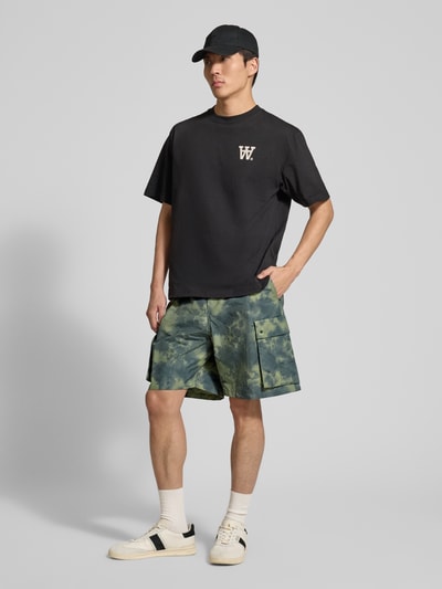 Männermodel trägt Shorts mit Cargotaschen Modell 'Ethan' von Wood Wood aus Baumwoll-Mix im Relaxed-Fit, grün gemustert, Ganzkörperansicht von vorne. Bundweite bei Größe M: 41 cm, Innenbeinlänge bei Größe M: 20 cm.