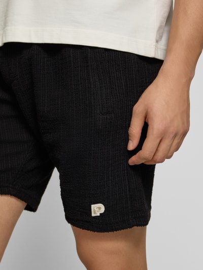 Männermodel trägt Shorts mit Label-Patch Modell 'LIBCO' von Pegador in Schwarz mit Strukturmuster, Material-Mix, Nahaufnahme Oberschenkel.