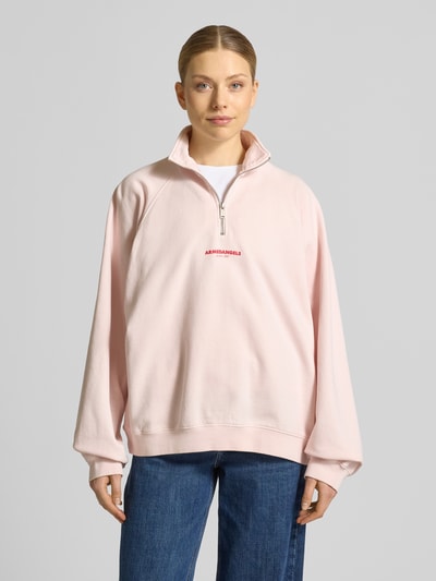 Frauenmodel trägt Oversized Sweatshirt aus reiner Baumwolle von Armedangels in Optik: Unifarben, Troyer-Kragen, Label-Print, Langarm, Raglanärmel, Materialverarbeitung: Sweat, Farbe hellrosa, Ganzkörperansicht von vorne.