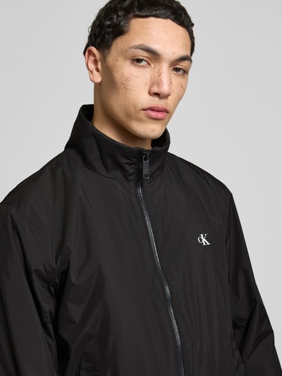 Mannenmodel draagt 'Trainingsjacke mit Logo und Stehkragen' van Calvin Klein Jeans in zwart, regular fit, staande kraag, logo, rits, steekzakken, halve close-up bovenlichaam.