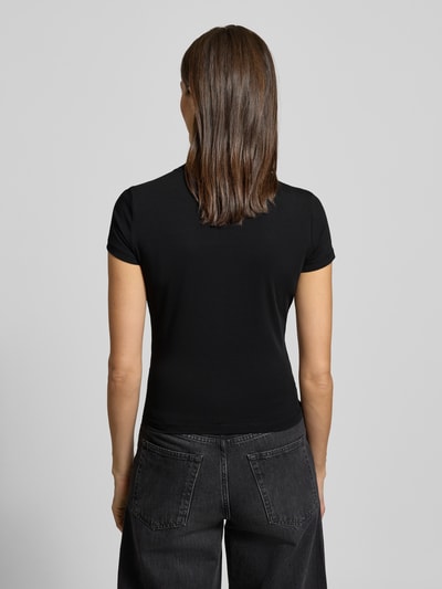 Kobieta modelka nosi Slim Fit T-Shirt mit Allover-Strasssteinbesatz od Guess, damski t-shirt, stójka, krój slim fit, obszycie z kamieni stras na całej powierzchni, detal z logo, mieszanka bawełny i elastanu, długość tyłu 51 cm dla rozmiaru S, czarny kolor, gładka faktura, widok całej sylwetki z tyłu.