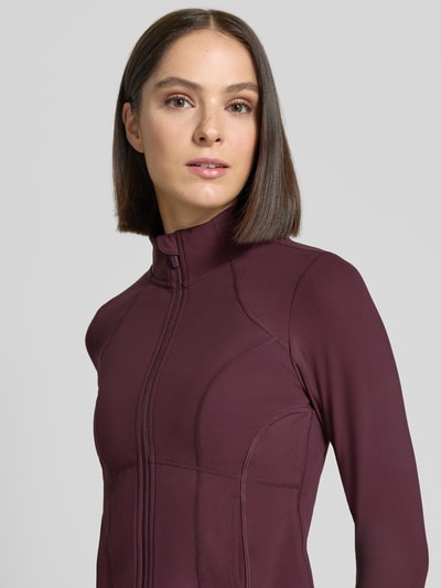 Vrouwenmodel draagt Slim Fit jack met opstaande kraag Model 'STORM' van Fabletics, kleur bordeaux, detailopname bovenlichaam. Ruglengte maat S: 45 cm, schouderbreedte: 35 cm, borstwijdte: 42 cm, mouwlengte: 68 cm, ritssluiting.
