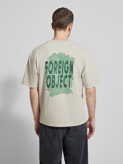 Męski model ma na sobie T-Shirt mit Logo-Print Modell 'Gary' od Wood Wood z bawełny, prosty krój, logo z tyłu, kolor beżowy, widok całej sylwetki od tyłu.