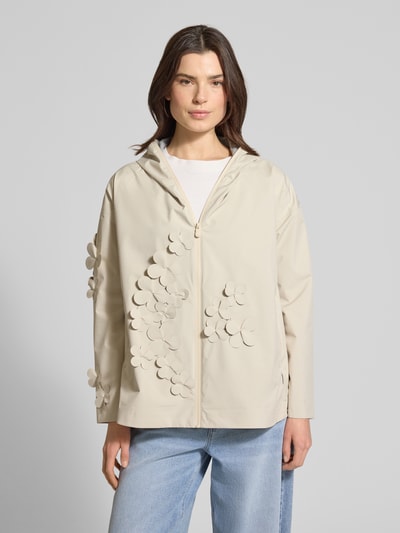 Frauenmodel trägt Regular Fit Regenjacke mit floralen Applikationen Modell 'Rosa' von SAVE THE DUCK in Beige, Kapuze, Logo-Patch, Ganzkörperansicht von vorne, Damenjacke, Reißverschluss, Rückenlänge 72 cm, Schulterbreite 57 cm, Brustweite 45 cm, Ärmellänge 50 cm.