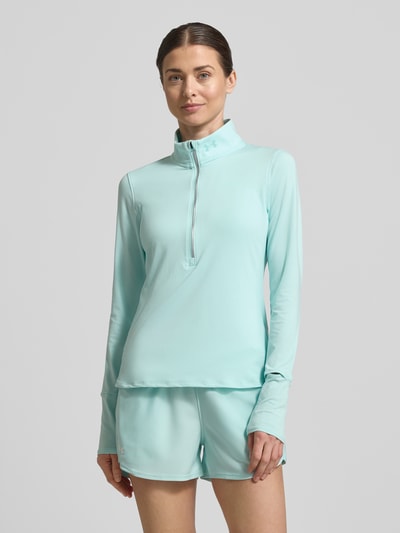 Frauenmodel trägt Longsleeve mit Stehkragen und Reißverschluss von Under Armour in Hellblau, glatte Oberfläche, Regular Fit, Ganzkörperansicht von vorne.