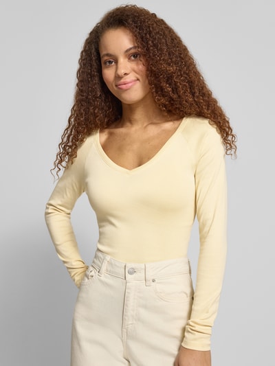 Modelka kobieca nosi Slim Fit Longsleeve mit V-Ausschnitt od Gina Tricot z elastycznej bawełny, krój slim fit, długi rękaw, kremowy kolor, widok z przodu, zbliżenie górnej części ciała.