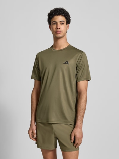 Männermodel trägt T-Shirt mit Label-Print von adidas Training in Olivgrün, Lockerer-Schnitt, gerippter Rundhalsausschnitt, Nahaufnahme Oberkörper. Rückenlänge bei Größe M: 70 cm.