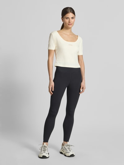 Frauenmodel trägt Cropped T-Shirt aus Lyocell-Mix Modell 'Circe' von Guess Activewear in Creme mit Logo-Stitching, Ganzkörperansicht von vorne.
