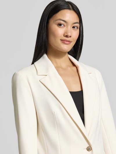 Frauenmodel trägt Slim Fit Blazer mit Stretch-Anteil Modell 'CLAUDIE' von GUESS in Creme mit Reverskragen und Strass-Knopf, Nahaufnahme Oberkörper.