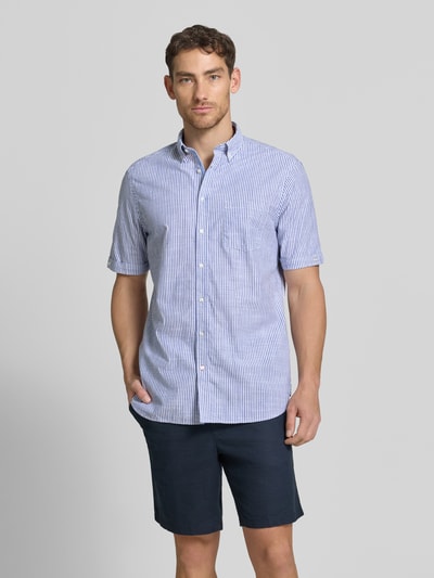 Męski model nosi Regular Fit Freizeithemd mit Brusttasche od Christian Berg Men z bawełny, w niebiesko-białe paski, kołnierzyk typu button down, krótki rękaw, listwa guzikowa, brustasza, długość tyłu 79 cm, szerokość w rozmiarze M 47 cm, długość rękawów 26 cm; zdjęcie całej sylwetki z przodu.