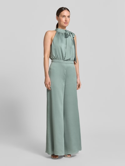 Modelka kobieca nosi Jumpsuit mit Schluppe und rückseitigem Knopfverschluss od Swing, satynowy, krój slim fit, kolor zielony, widoczne wiązanie przy szyi, szerokie nogawki, Szerokość w rozmiarze 36: 66 cm, długość tyłu: 37 cm, długość całkowita: 152 cm, długość nogawki wewnętrznej: 82 cm; ujęcie całej sylwetki z przodu